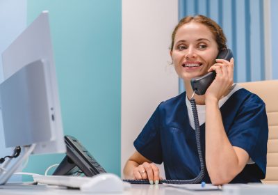 smiling-young-woman-nurse-receptionist-talking-on-phone-while-working-in-modern-clinic